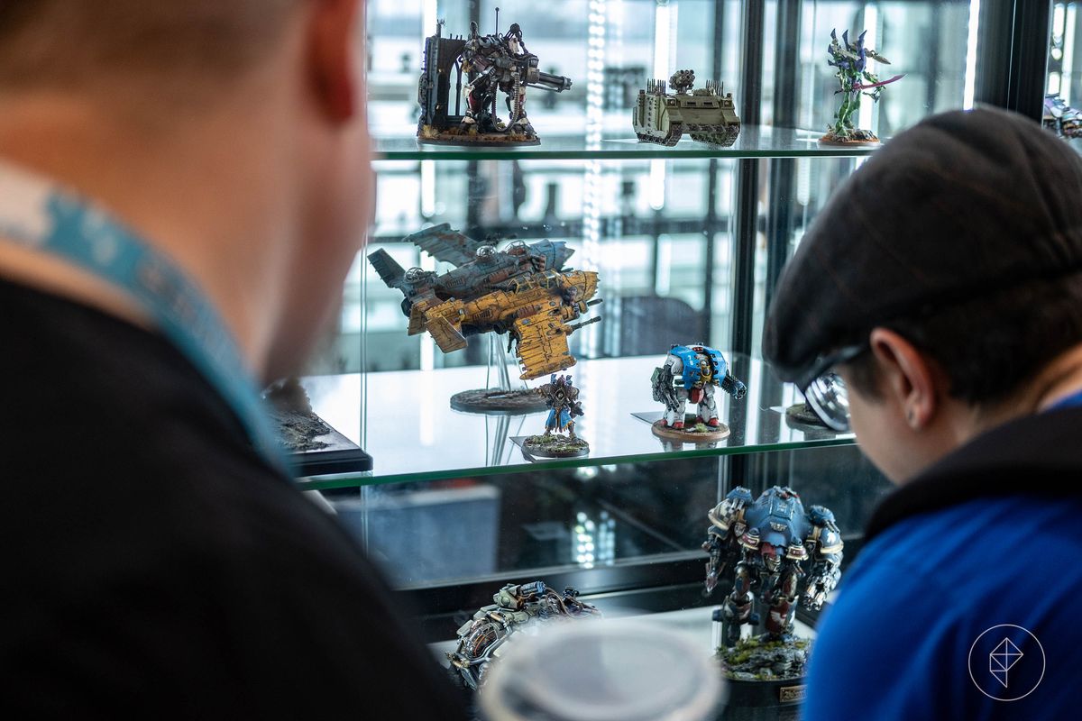 A half-dozen finished pieces at AdeptiCon 2023, including a ship, a robot, and a tank, arranged in a display case. A central miniature is an ork plane, with two fuselages fused together into a singular monstrosity. Below, a large knight strides forward with its shoulder lowered, like a linebacker blitzing the quarterback.