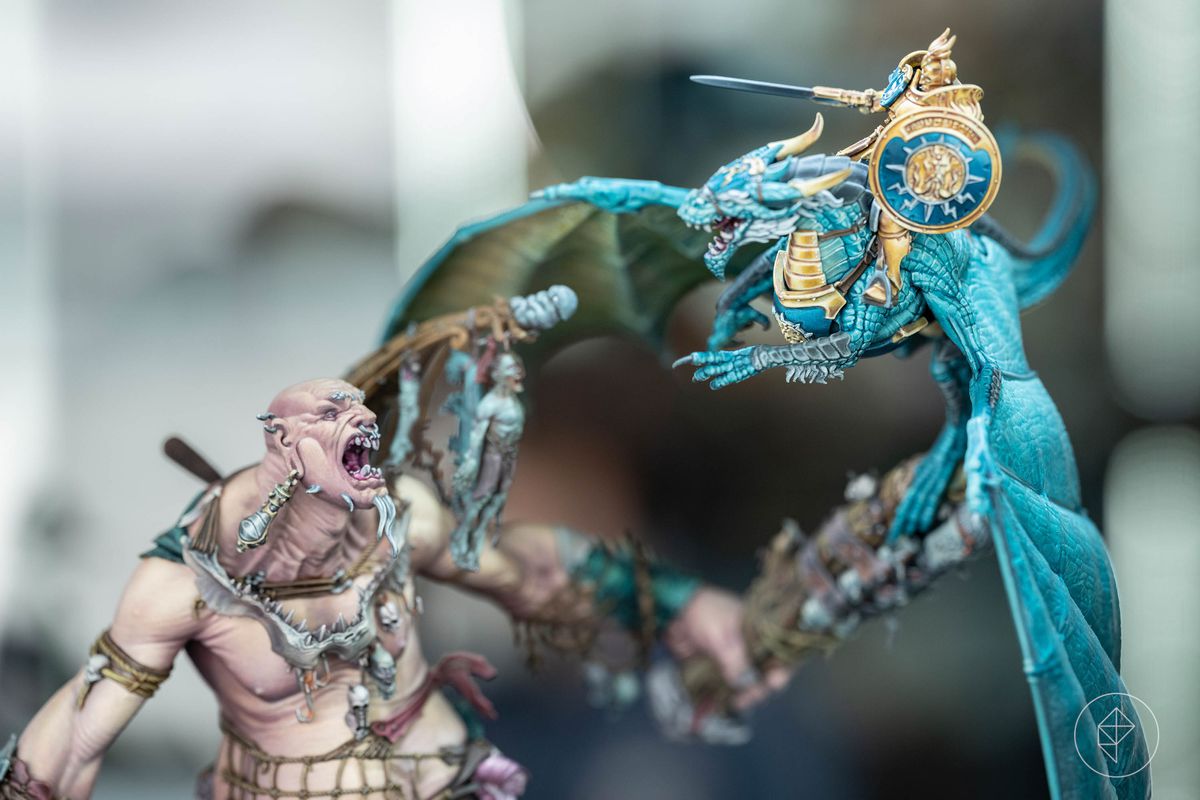 A mega-gargant with pale, pinkish skin and a Sigmarite on a blue drake fight in a duel at the Golden Demon 2023. Both miniatures show their details differently, the mega-gargant its folds of taught skin and the drake its many thousands of scales highlighted in white paint.