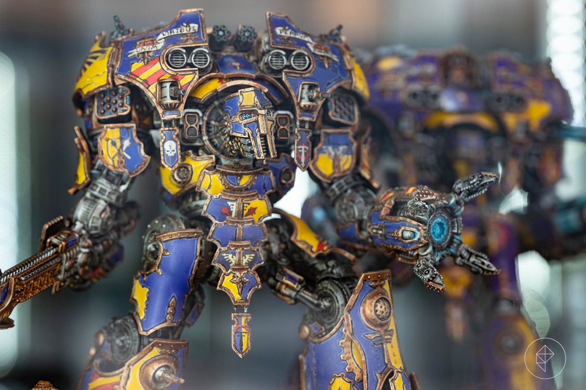 A massive titan, rendered in fractional scale for the Adeptus Titanicus game. It is elaborately painted all over with alternating blue and yellow panels, with rust and wear and energy coursing through its weapons. It head is up, its eyes looking at the viewer even as its gigantic guns traverse to meet its gaze.