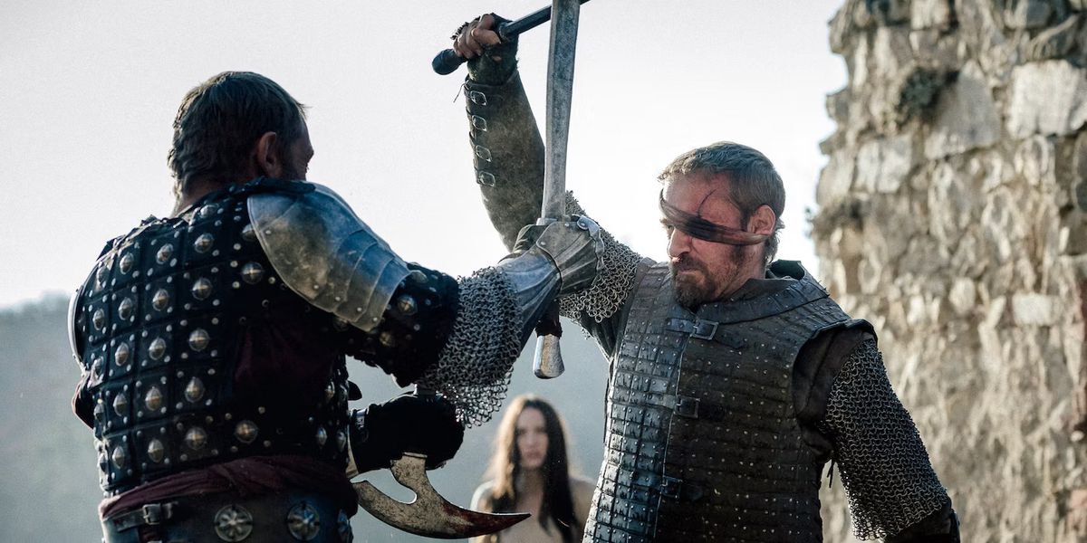 Un uomo con una sciarpa insanguinata sopra l'occhio sinistro (Ben Foster) in armatura medievale si scontra con le spade con un avversario corazzato con una giovane donna che guarda sullo sfondo.
