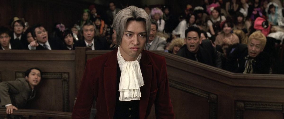 La folla nell'adattamento live action di Ace Attorney risponde sporgendosi in un momento cruciale, dietro il volto accigliato di Miles Edgworth.