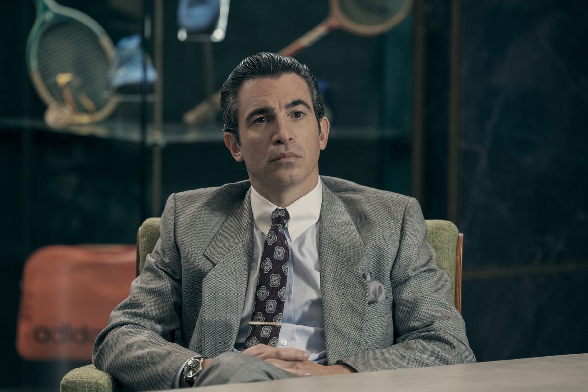 Chris Messina nei panni di David Falk siede in una sala riunioni aziendale con un abito grigio nel film Air.