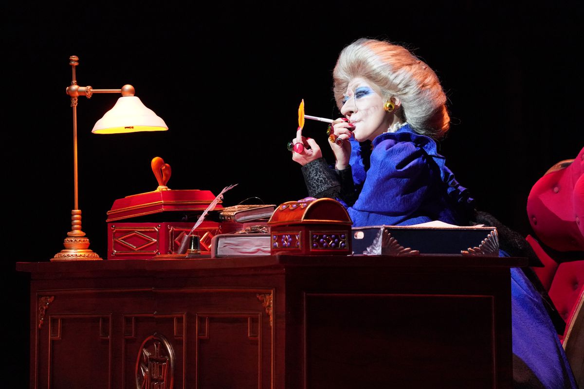 La strega Yubaba, con un vestito blu e un esagerato sbuffo di capelli grigi, siede a una scrivania e accende una sigaretta con una fiamma che le sporge dal dito in La città incantata: Live on Stage