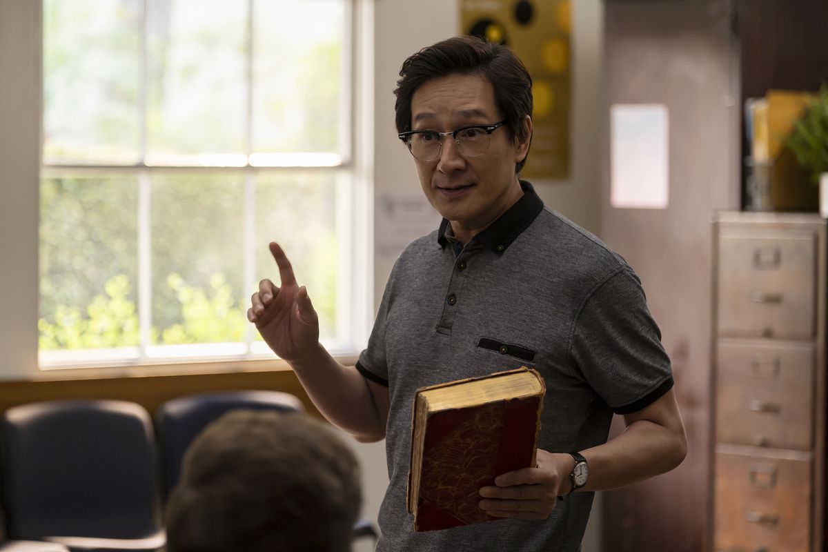 Ke Huy Quan in cinese nato americano.  Tiene in mano un libro e sembra che stia insegnando davanti a una classe.