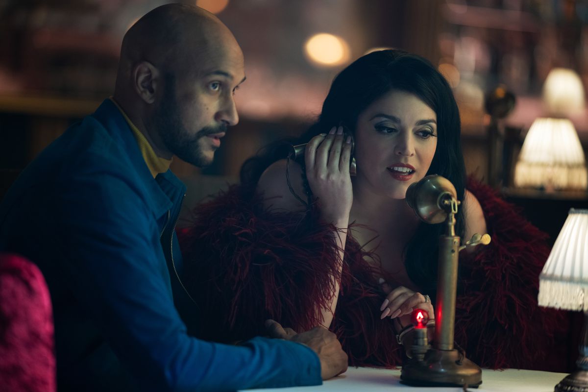 Melissa (Cecily Strong) usa un telefono d'altri tempi a un tavolo mentre Josh (Keegan Michael-Key) si siede accanto a lei guardando qualcosa in un fotogramma della seconda stagione di Schmigadoon