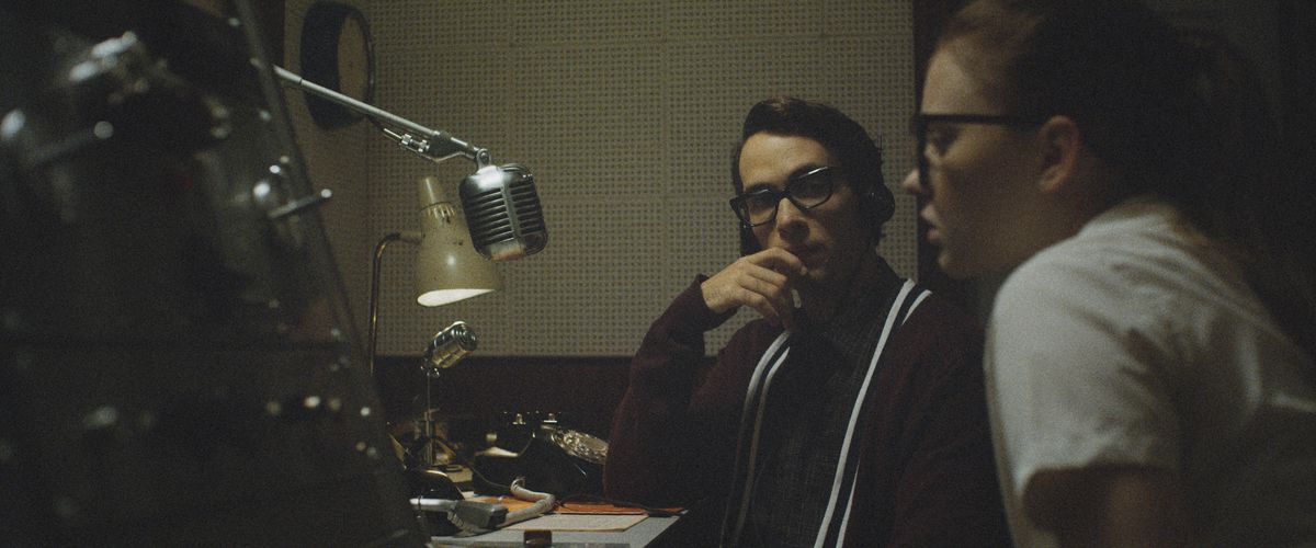 A young man and woman, both in heavy 1950s glasses, sit at a counter in front of an old-school heavy radio mic in The Vast of Night.