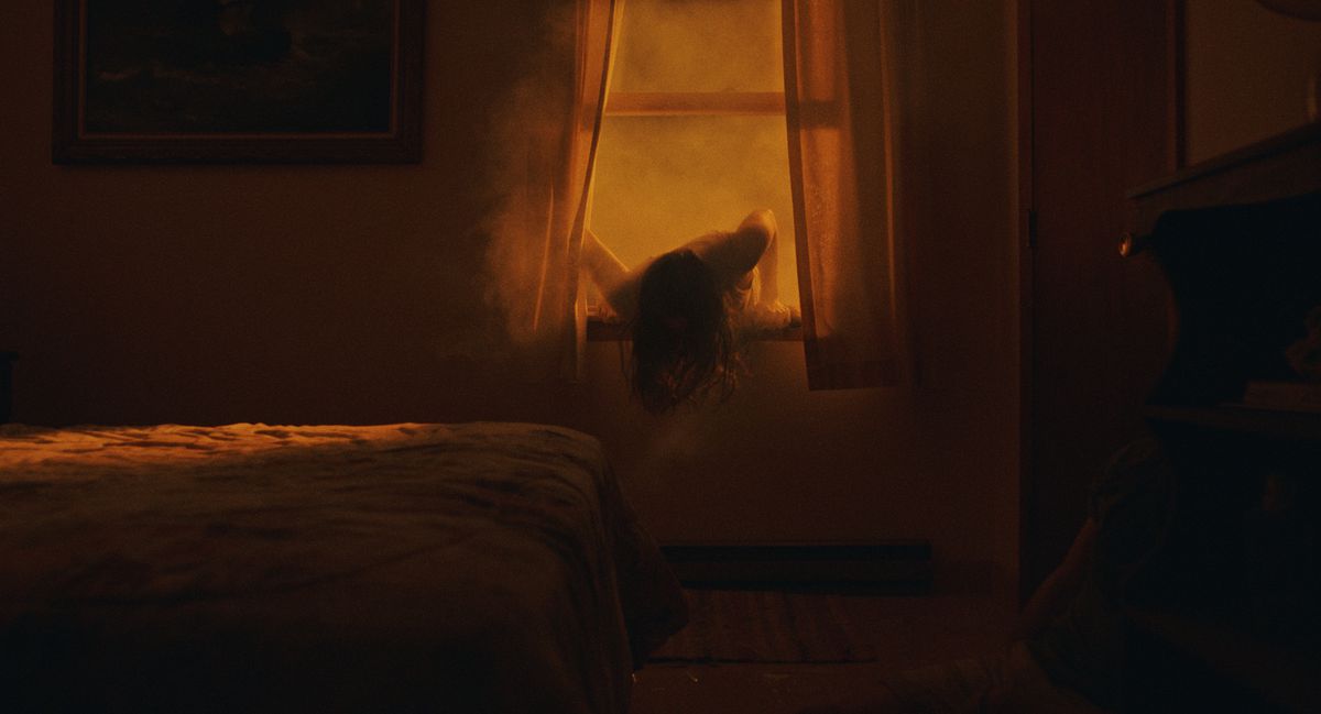 Una donna dai capelli scuri striscia attraverso una finestra aperta in una stanza illuminata da una misteriosa luce giallo arancione in The Beach House.