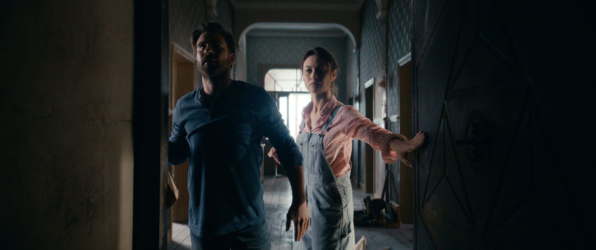 Olga Kurylenko e Kevin Janssens esplorano un corridoio buio della loro casa nel film horror The Room. 
