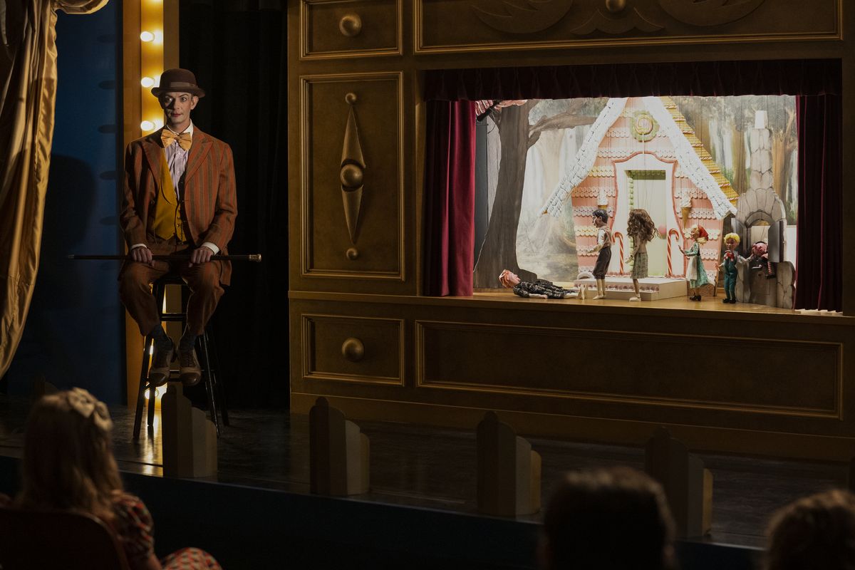 Un uomo con un trucco da vaudeville siede su un palco con uno spettacolo di marionette illuminato in Ratched