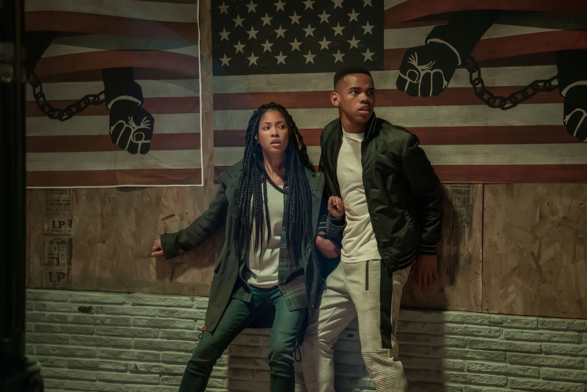 Nya (LEX SCOTT DAVIS) and Isaiah (JOIVAN WADE) stand in front of chained up arms and the american flag in the first purge