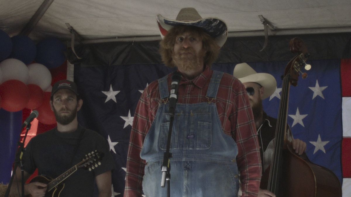 Sacha Baron Cohen, disguised as a country singer, stands on a stage in overalls and a cowboy hat in Borat Subsequent Moviefilm