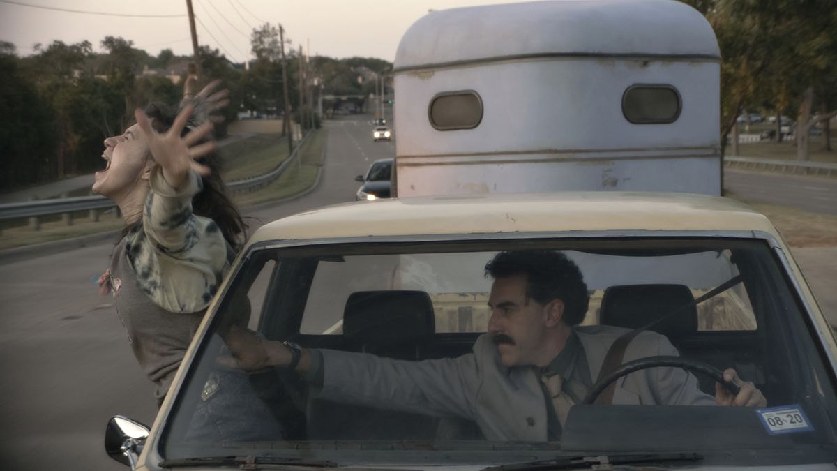 Maria Bakalova leans out of the window of a moving car as Sacha Baron Cohen tries to pull her back in in Borat Subsequent Moviefilm