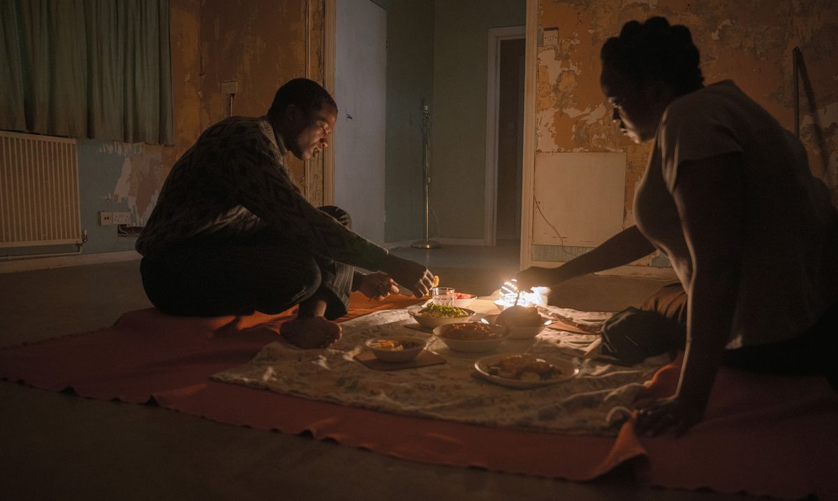I rifugiati sudanesi Bol (Sope Dirisu) e Rial (Wunmi Mosaku) cenano sul pavimento della loro casa a schiera non arredata (e molto infestata) nella sua casa.