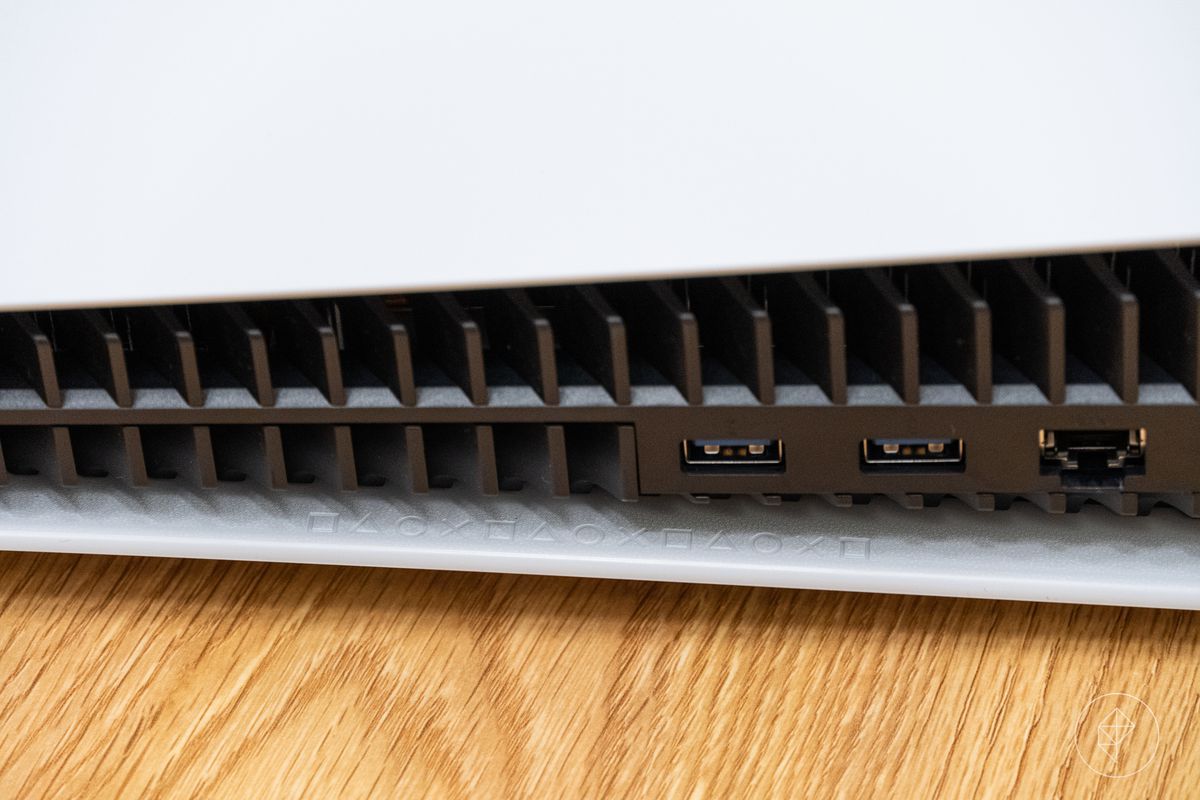 un primo piano delle icone dei pulsanti frontali della PlayStation incise all'interno di uno dei pannelli laterali bianchi della PlayStation 5, appena sotto una delle porte USB 3.0 della console