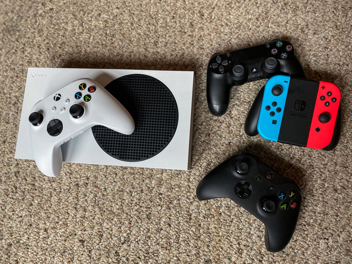 an overhead photo of the Xbox Series S console sitting on beige carpet with an Xbox controller on top of it, next to a black PlayStation 4 DualShock 4 controller; a Nintendo Switch Grip Controller with blue and red Joy-Cons in it; and a black Xbox One controller