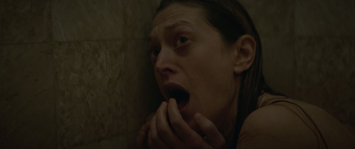 Una donna bagnata, curva sotto la doccia, urla, guardando qualcosa fuori dallo schermo