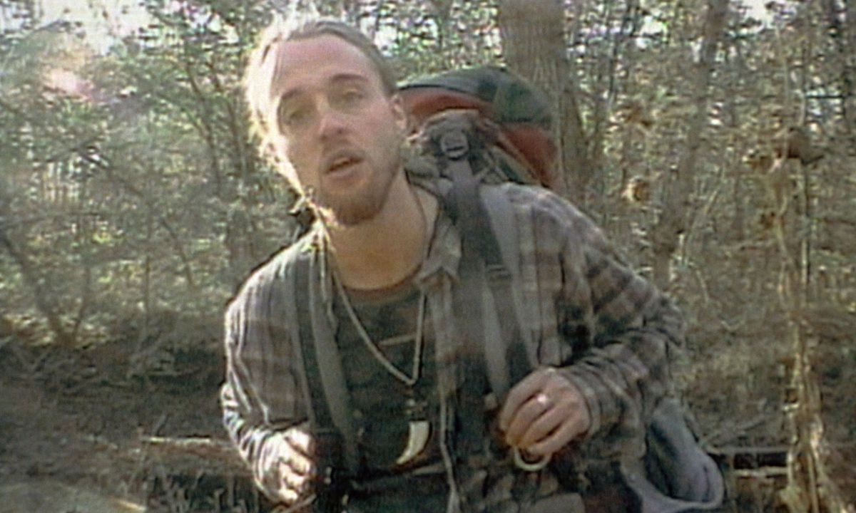 Joshua Leonard, in una sporca flanella e uno zaino pesante, è in piedi in una foresta a bocca aperta davanti alla telecamera in The Blair Witch Project.