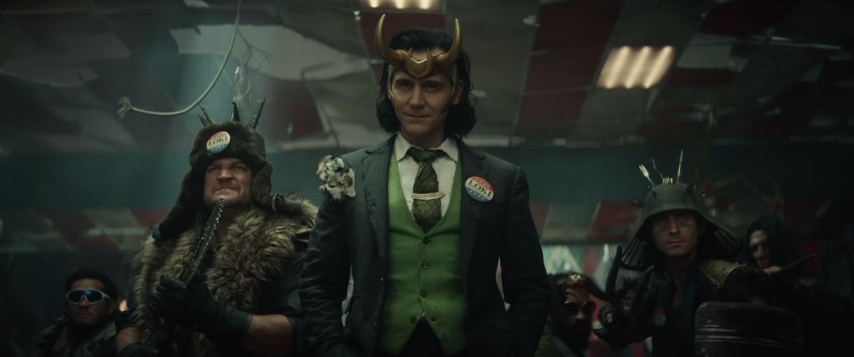 Tom Hiddleston nei panni di Loki, vestito con un abito lacero con una spilla VOTE LOKI, a Loki (Disney Plus)