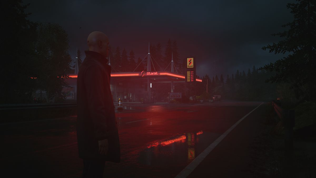 Agent 47 wearing a long coat with his collar popped, standing on a lonely road in a forested area at night with a gas station in the background in Hitman 3