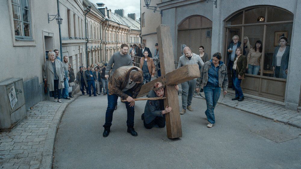 An exhausted man with a crown of thorns carries a giant wooden cross down a narrow street as a crowd in blue jeans follows close behind.
