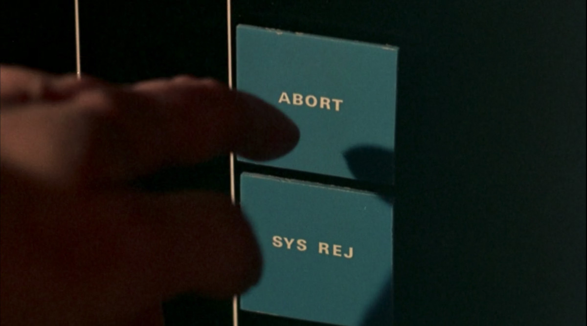 Close-up of a finger reaching to touch the “abort” button on an abortion machine in ZPG