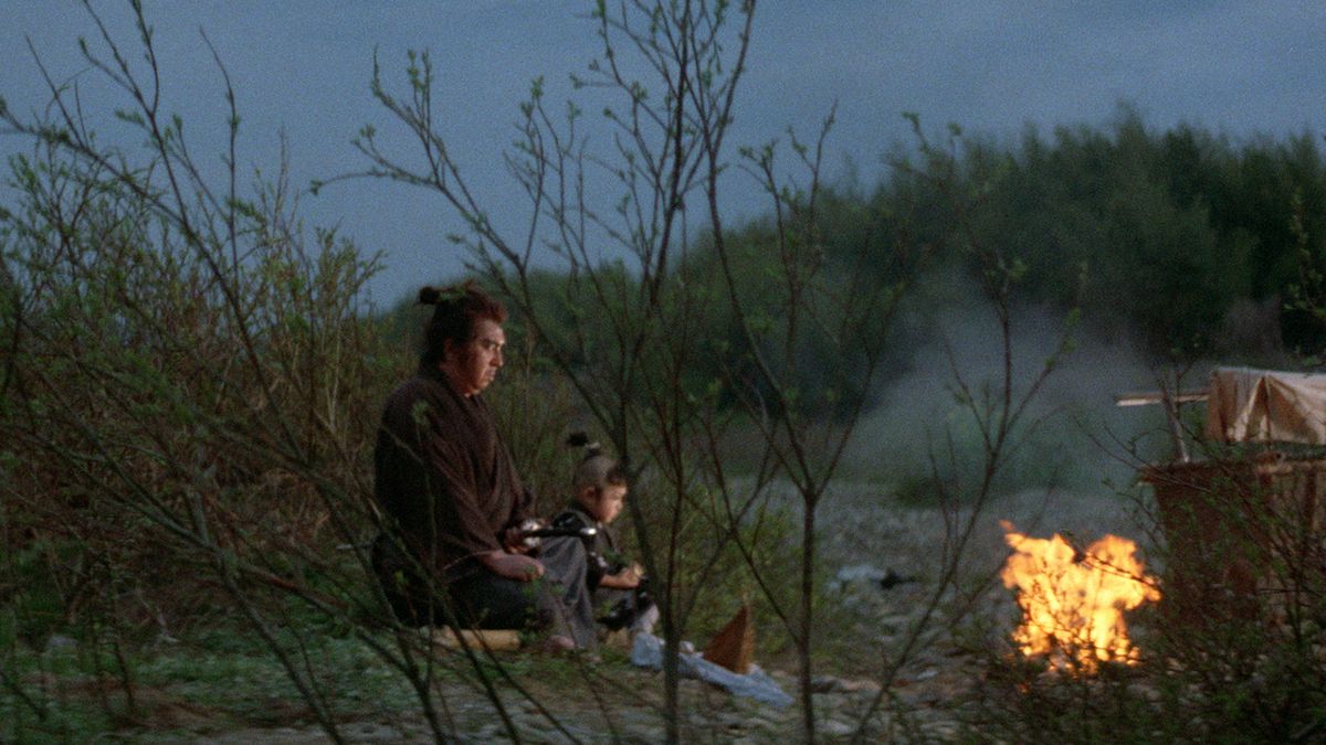 Un samurai in abito marrone con un taglio di capelli nodoso (Tomisaburo Wakayama) siede accanto a un fuoco con suo figlio (Akihiro Tomikawa) in un campo di canne.