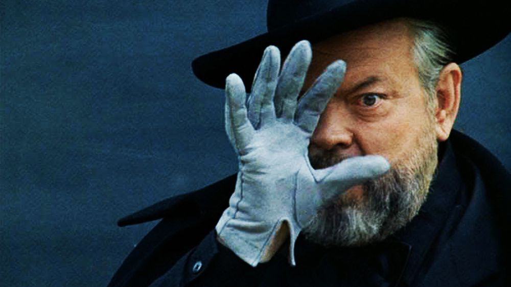 Un uomo barbuto (Orson Welles) che indossa un cappello nero, un cappotto e un guanto bianco che tiene la mano destra contro il viso.
