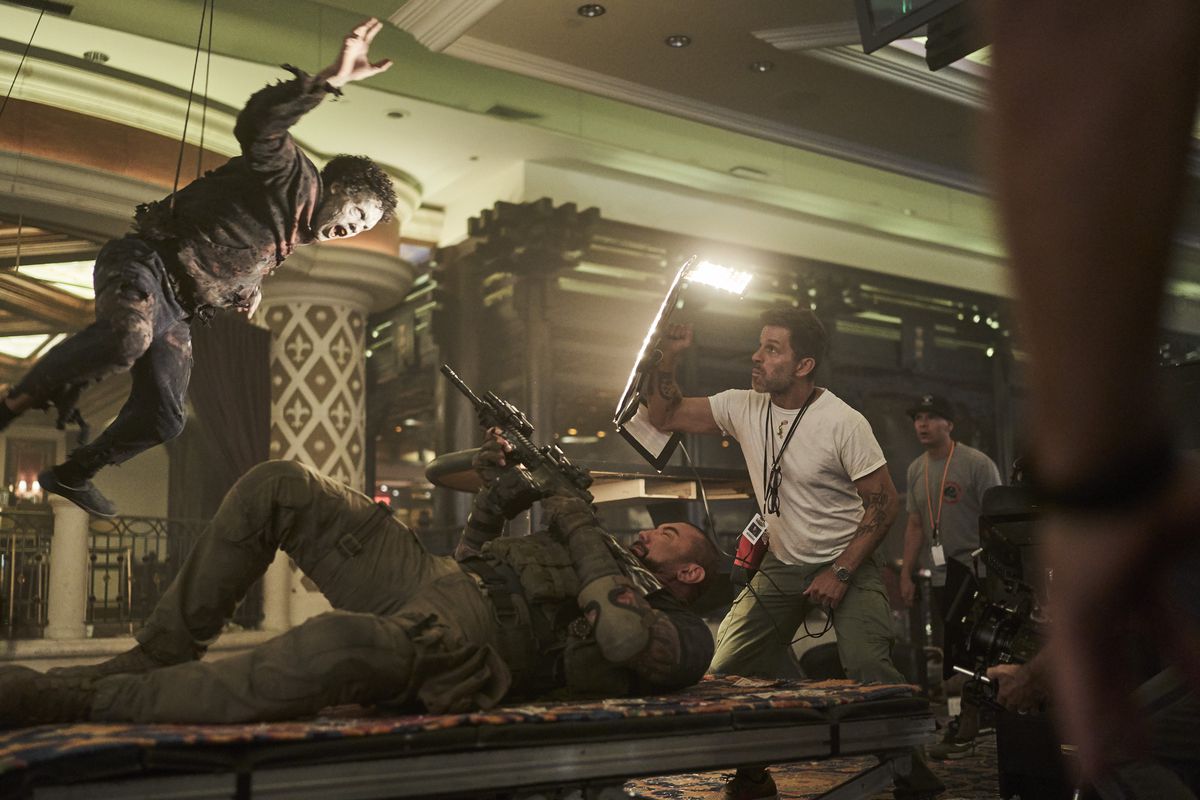 Dave Bautista giace supino, puntando la sua pistola verso un attore zombi sospeso sopra di lui su fili, mentre il regista Zack Snyder tiene un riflettore di luce sopra di lui sul set di Army of the Dead