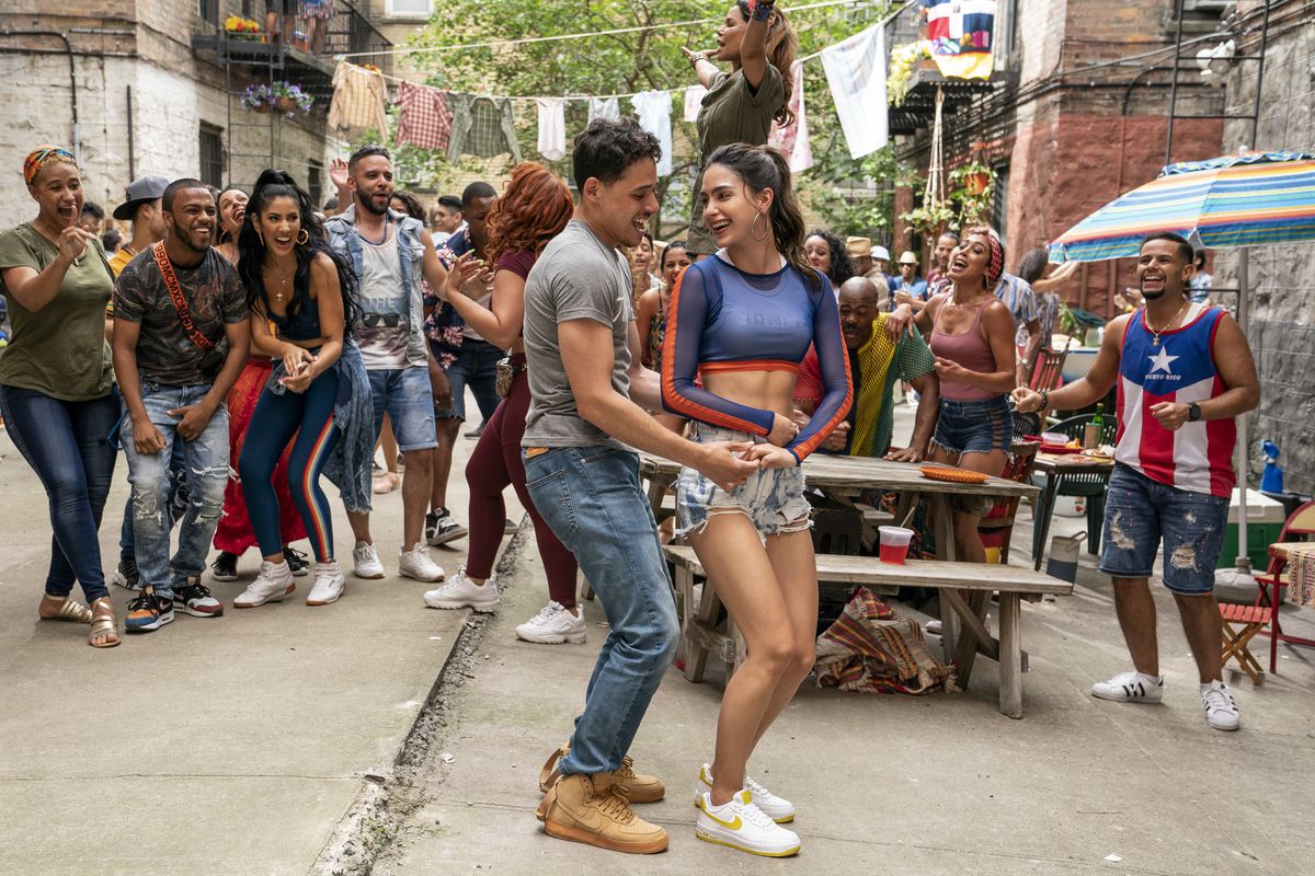 un uomo e una donna ballano davanti a un tavolo da picnic mentre i loro amici li incoraggiano in In the Heights