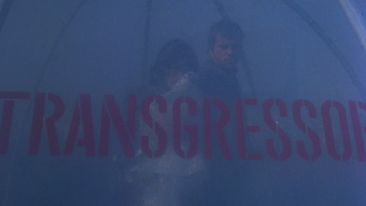 The stars of ZPG are dimly visible behind a huge stenciled sign that reads TRANSGRESSION in red