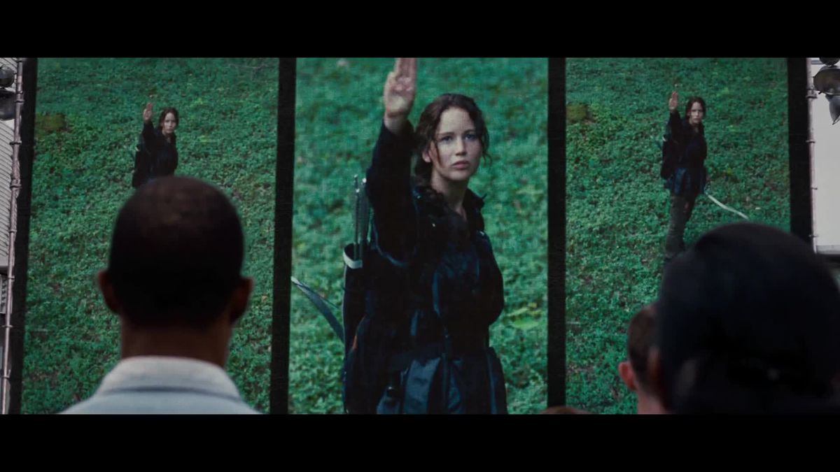 Diverse persone viste da dietro guardano una serie di monitor mentre Katniss Everdeen dà il segno con la mano del Ghiandaia Mocking in The Hunger Games