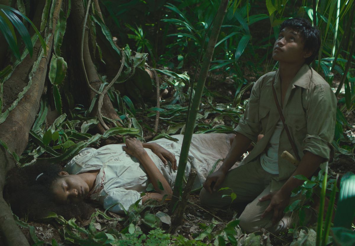 Una donna in bianco giace nella giungla mentre un giovane si inginocchia accanto a lei, guardando in alto
