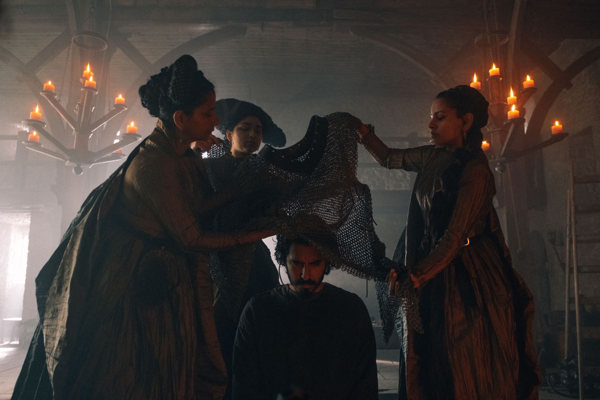 Tre donne abbassano una maglia a catena su un Dev Patel inginocchiato in The Green Knight di David Lowery