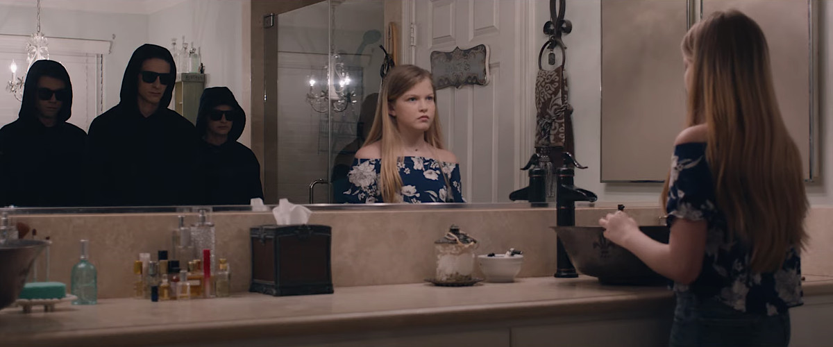 Tre adolescenti inquietanti in felpe scure e occhiali da sole stanno dietro una giovane ragazza bionda nel riflesso del suo specchio del bagno in Let Us In 