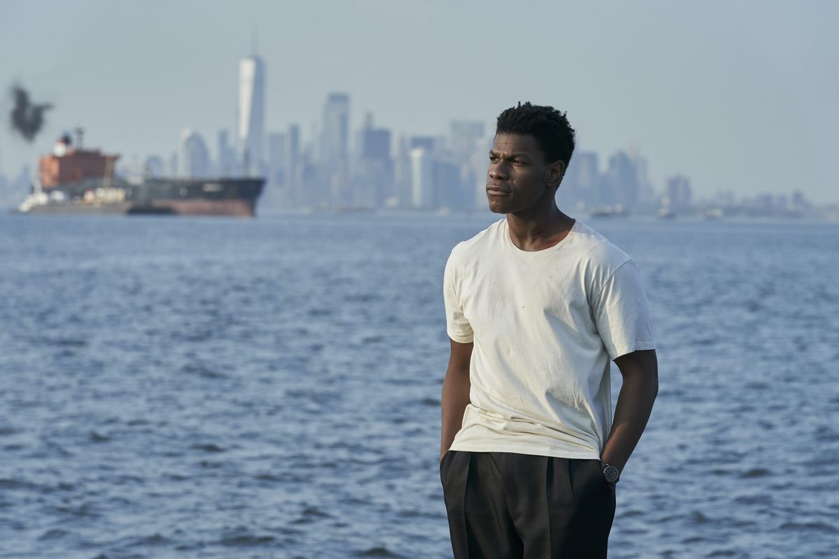 John Boyega, stravaccato con indosso una maglietta bianca e pantaloni larghi neri, sta vicino all'acqua con Manhattan sullo sfondo in Naked Singularity