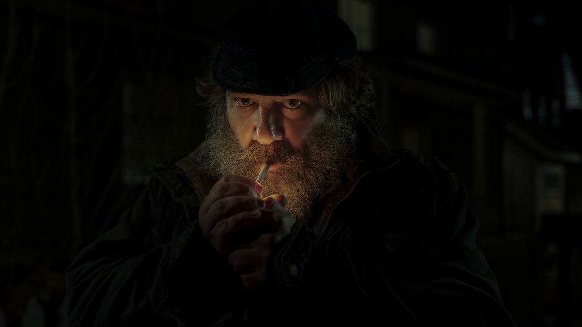 Un uomo con la barba pesante accende una sigaretta al buio, illuminandosi drammaticamente il viso, nella messa di mezzanotte