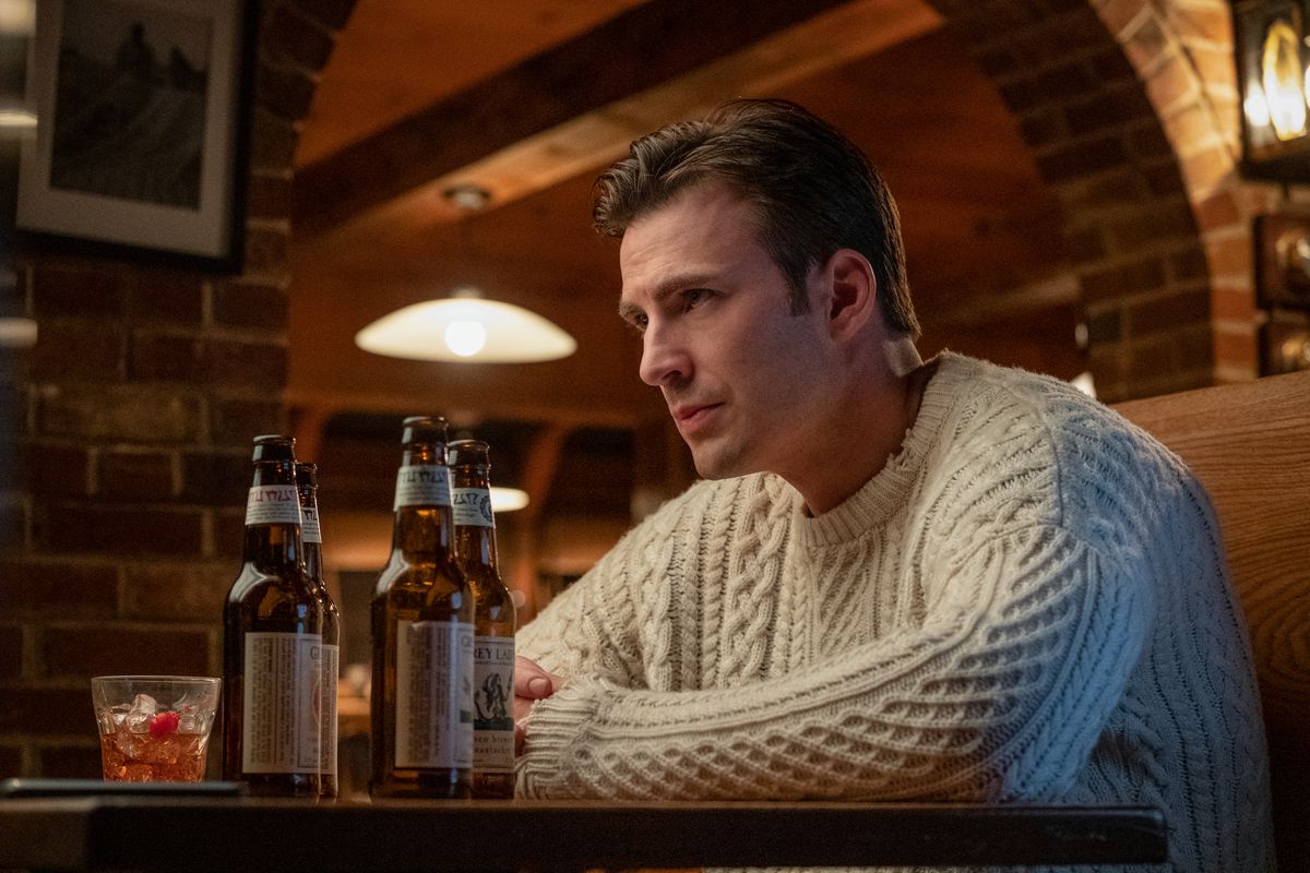 Chris Evans siede in un maglione con le braccia incrociate su un tavolo con due birre accanto a lui in Knives Out