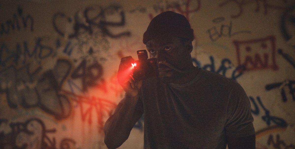 Yahya Abdul-Mateen II stands in a dark room, aiming a camera at graffiti in the 2021 Candyman