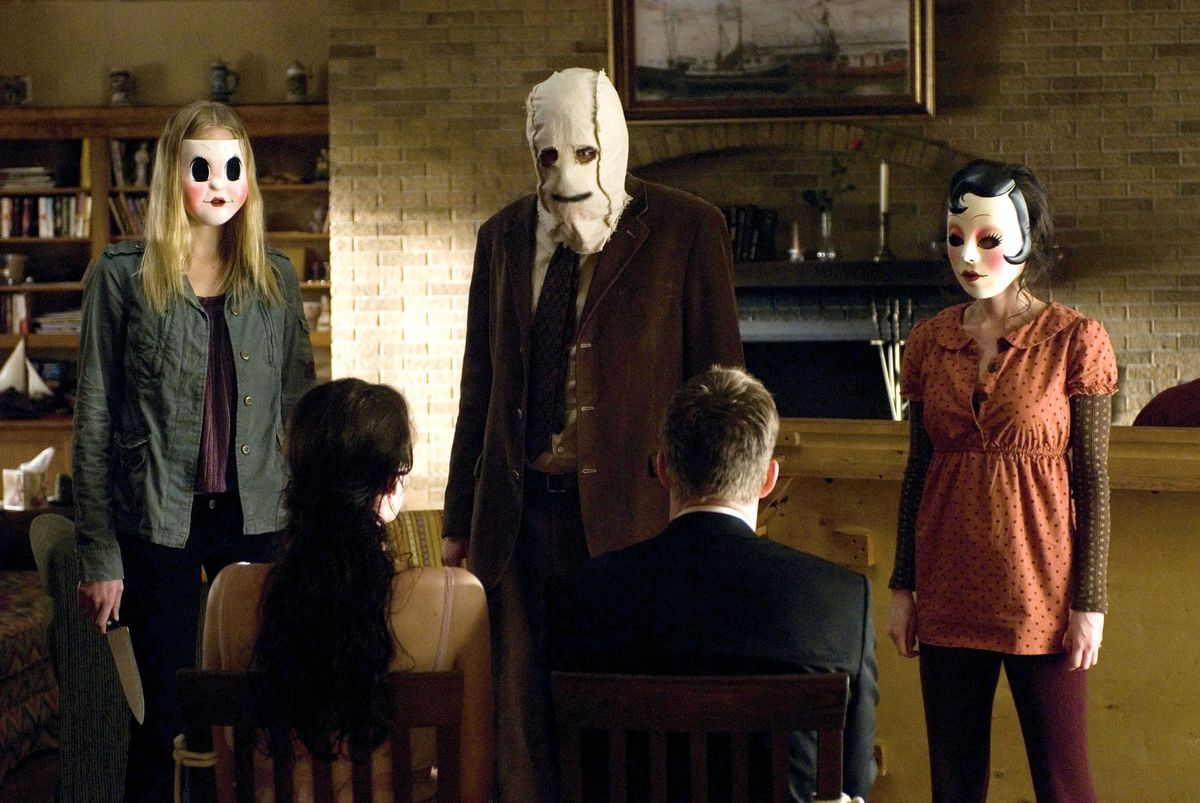 Three masked killers stand in front of a couple tied to chairs in their living room in The Strangers