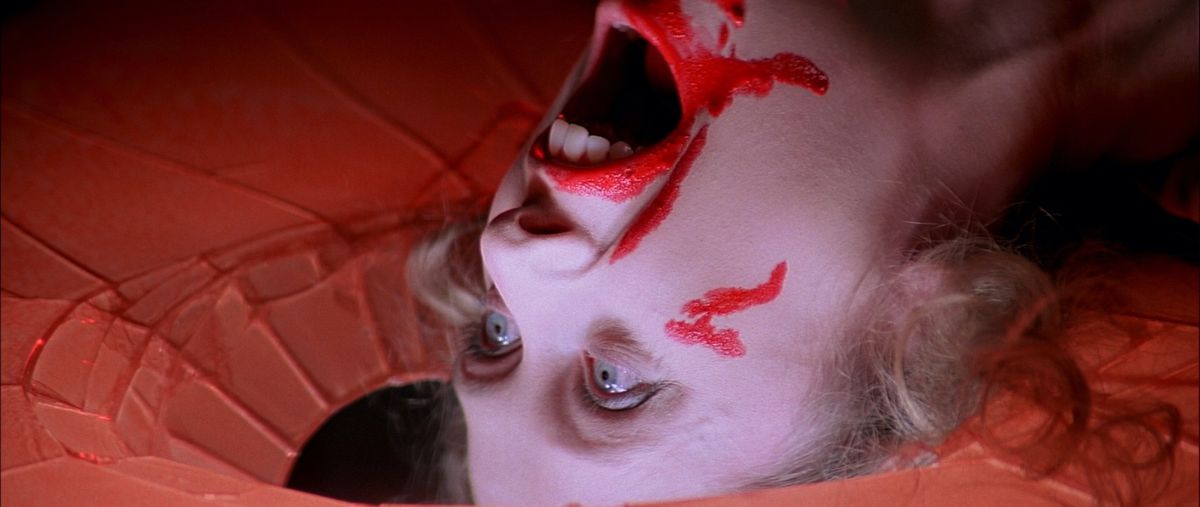 Una donna morta sdraiata con la testa su un pavimento in frantumi, sangue rosso neon intorno alla bocca, la testa gettata all'indietro in un ultimo grido, in Suspiria del 1977