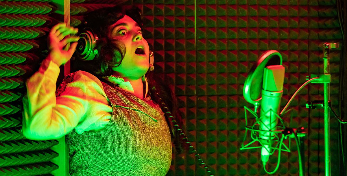 Una donna illuminata in modo vivido di verde e arancione nel Berberian Sound Studio, in piedi in una cabina di registrazione con un microfono argentato e pareti imbottite, con l'aria terrorizzata