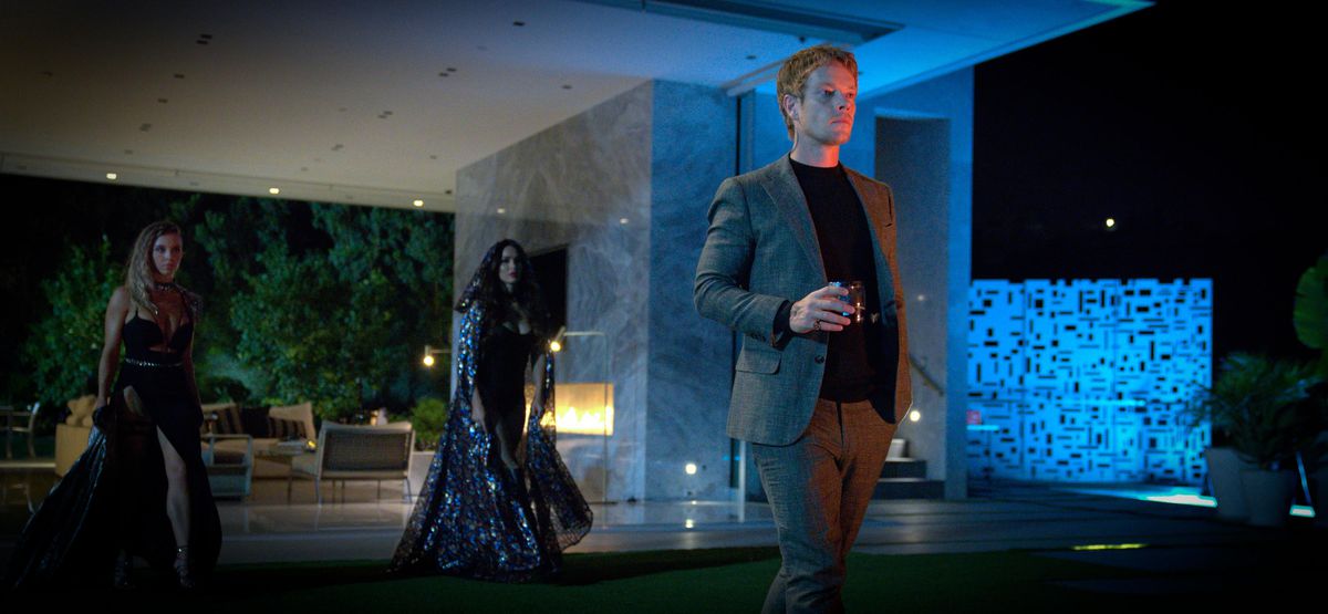 Alfie Allen tiene un drink e sta in piedi in un cortile dall'aspetto mod di notte con altri due vampiri dietro di lui che indossano abiti elaborati in Night Teeth