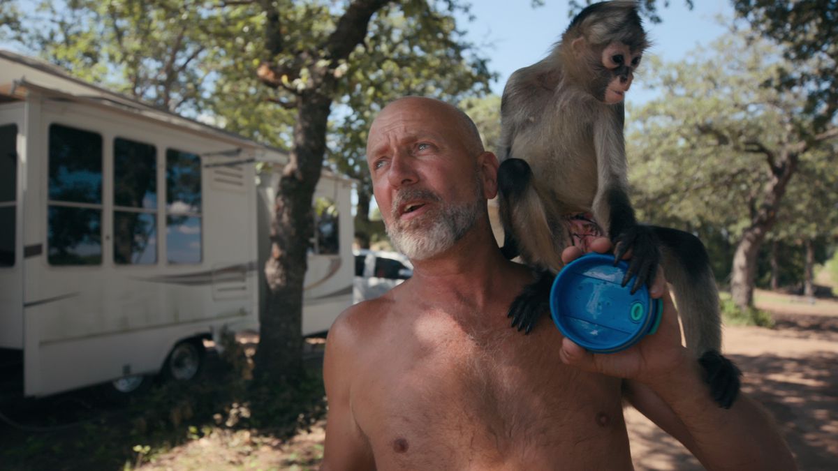 Il proprietario della fauna selvatica Tim Stark, a torso nudo e con una scimmia appollaiata sulla spalla, in piedi di fronte a una roulotte in Tiger King 2
