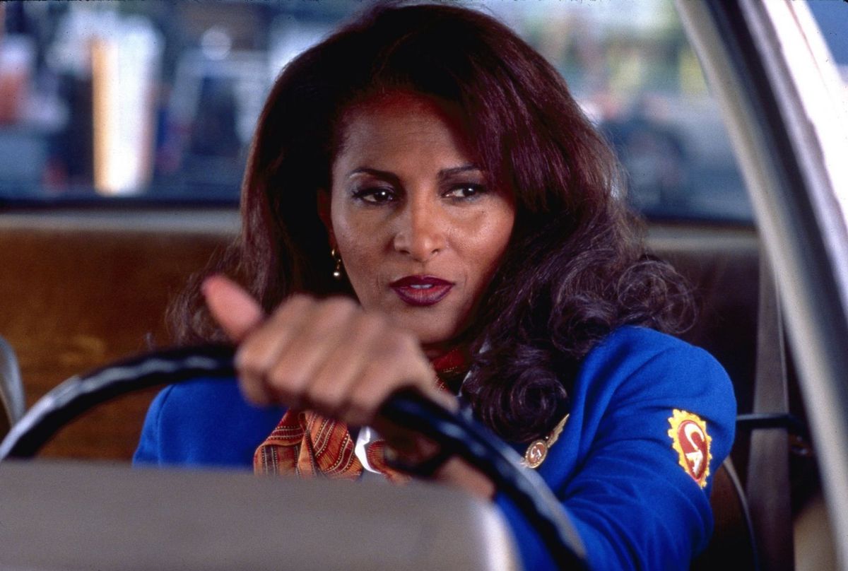 indossando una giacca blu da assistente di volo, Jackie (Pam Grier) siede al volante di un'auto in Jackie Brown