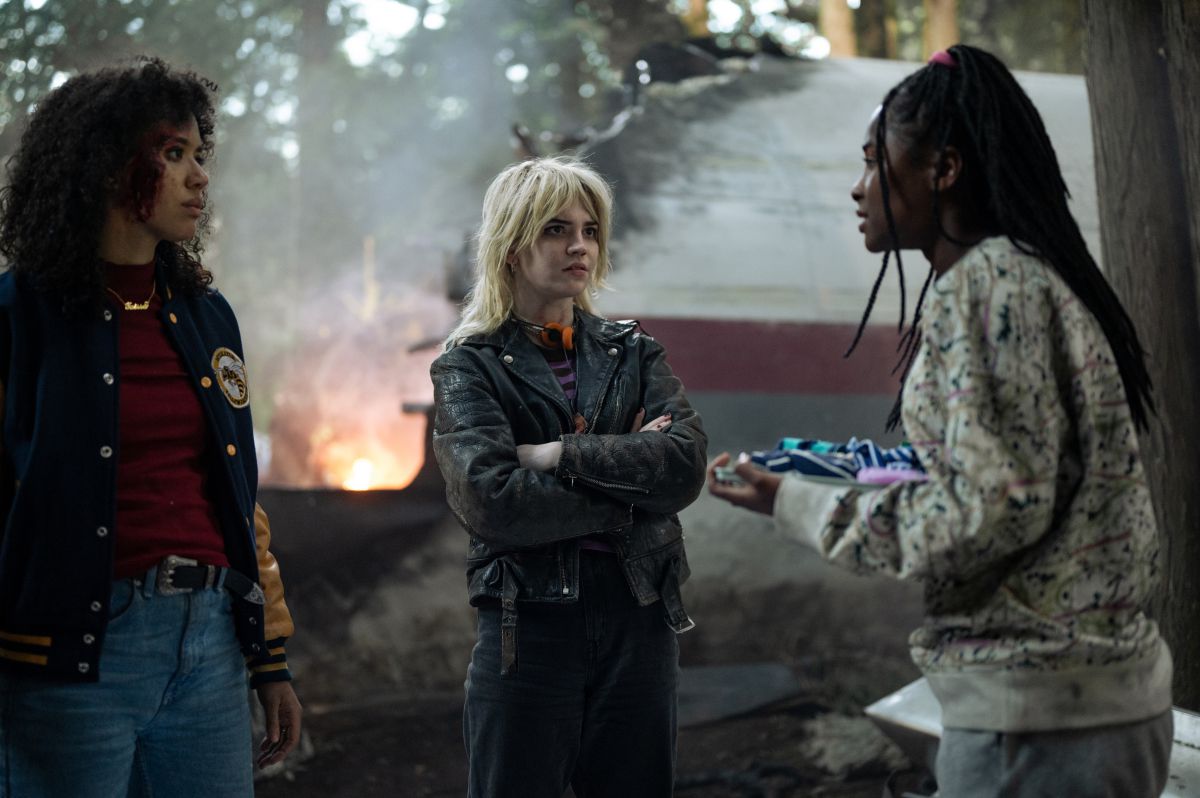 Three characters in Yellowjackets standing in front of a burning crashed plane in a still from the pilot