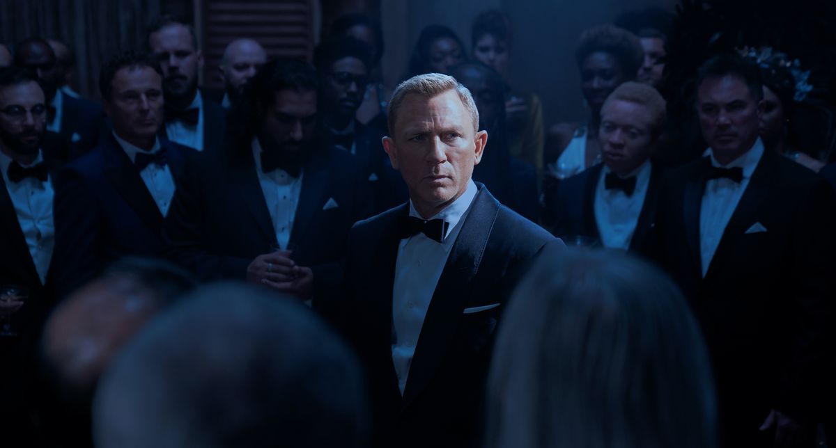 Daniel Craig nei panni di James Bond in No Time to Die, in piedi sotto i riflettori, circondato da membri in abito formale di SPECTRE