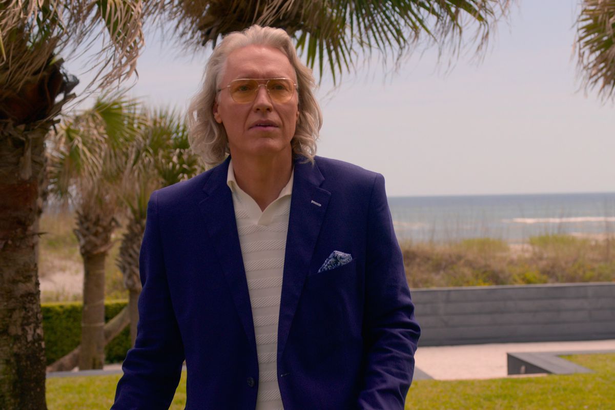 Un uomo con occhiali scuri e cerchiati di metallo e lunghi capelli grigi che indossa una giacca sportiva blu.  La spiaggia è in lontananza.