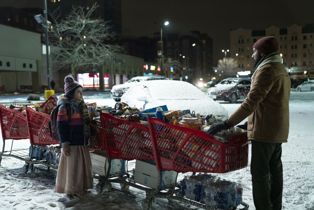 Kirsten (una ragazza) in piedi accanto a un certo numero di carrelli della spesa pieni di cibo, mentre Jeevan li trascina attraverso un parcheggio coperto di neve.