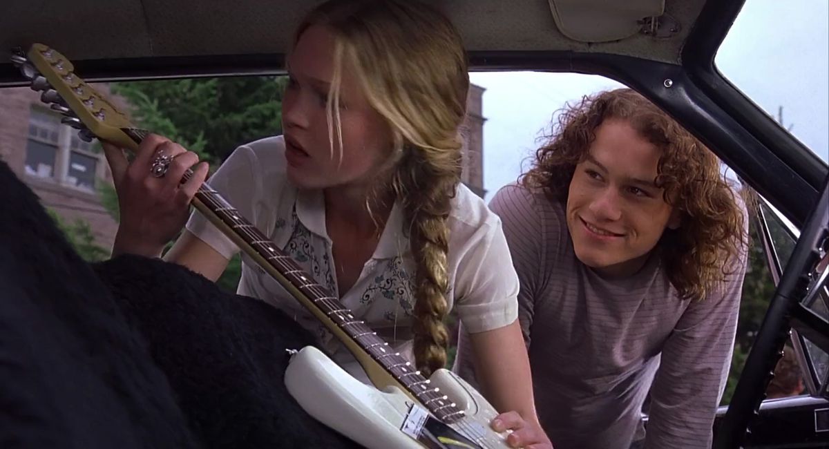 Julia Stiles reagisce con sorpresa a una chitarra bianca lasciata nella sua macchina da Heath Ledger, che guarda con un sorriso.