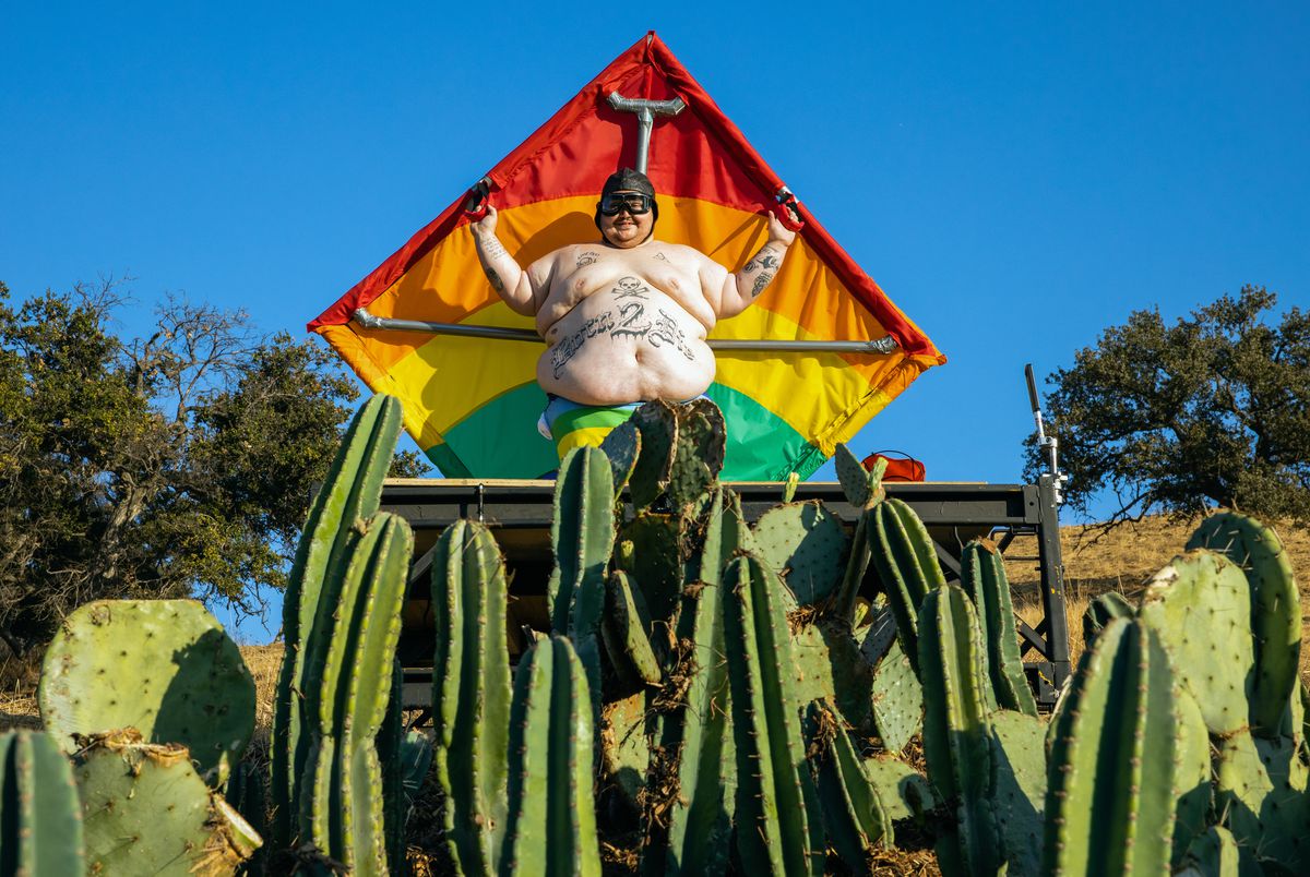 Zach Holmes, un uomo tatuato estremamente grande e senza maglietta, tiene in mano un deltaplano e sorride mentre si trova sopra un campo di cactus